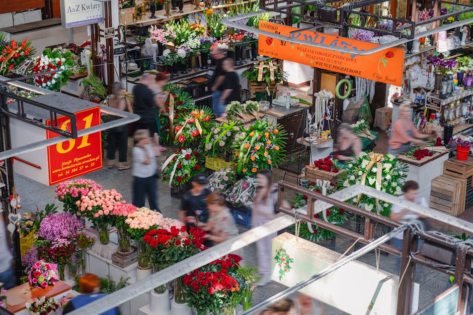 A bustling indoor flower market with multiple floral stalls displaying a variety of fresh flower arrangements. The scene includes bouquets of pink, red, purple, and white flowers such as chrysanthemum